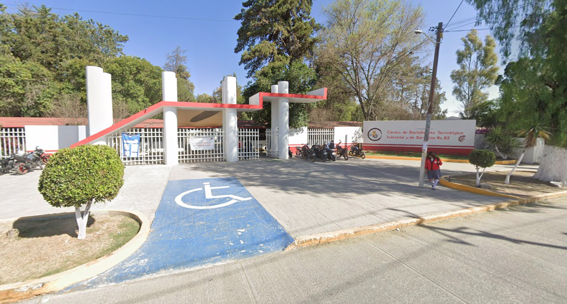 Fotografía en la que se muestra una explanada a la que se puede ingresar por una rampa. Al terminar la explanada hay un pequeño techo, sostenido por cuatro pilares. Detrás de los pilares hay una reja que da acceso al plantel y a la derecha de la reja hay un muro blanco que en letras de color naranja dice el nombre del plantel.